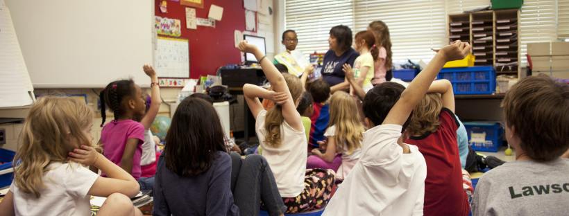 Stock photo of a classroom of children, probably aged about eight. They are sat on the floor, some with their hands up, while the teacher talks to three children in front of the group. I think these three children may be presenting. The classroom looks busy, lots of bright colours and artwork on display