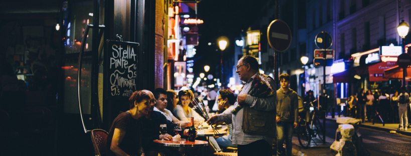 Stock photo of a busy Parisian street at night. People are sat at small round tables on the pavement and a chalkboard advertises Stellla at 5.5 a pint